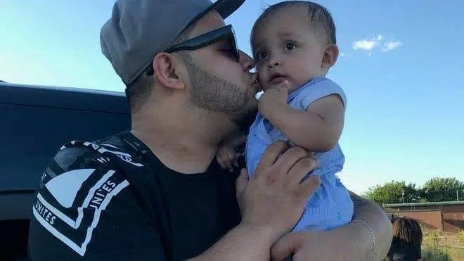 A man wearing sunglasses kisses a baby on the cheek while holding them in his arms.