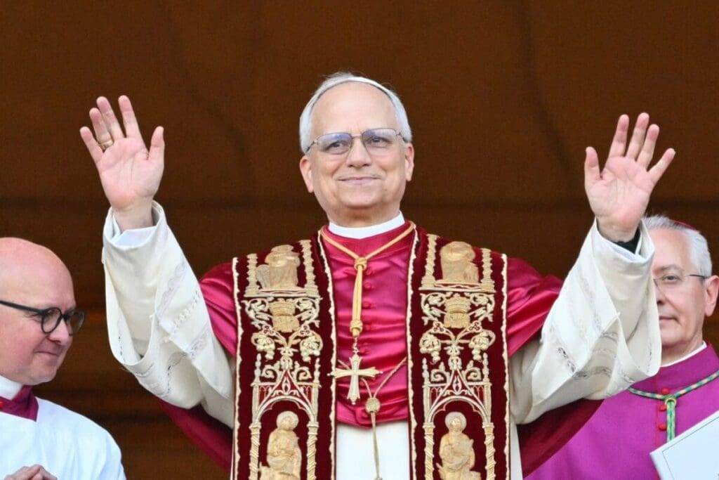 Cleric wearing ornate vestments and glasses, raising hands in a greeting gesture.